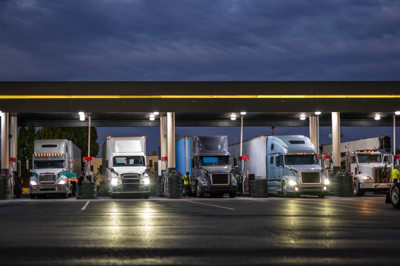 Semi trucks fueling at a lit truck stop at dusk, representing round-the-clock expedited freight operations