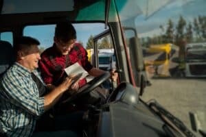 Truck driver and coordinator reviewing load paperwork in a semi cab, showing transportation back office documentation at the source