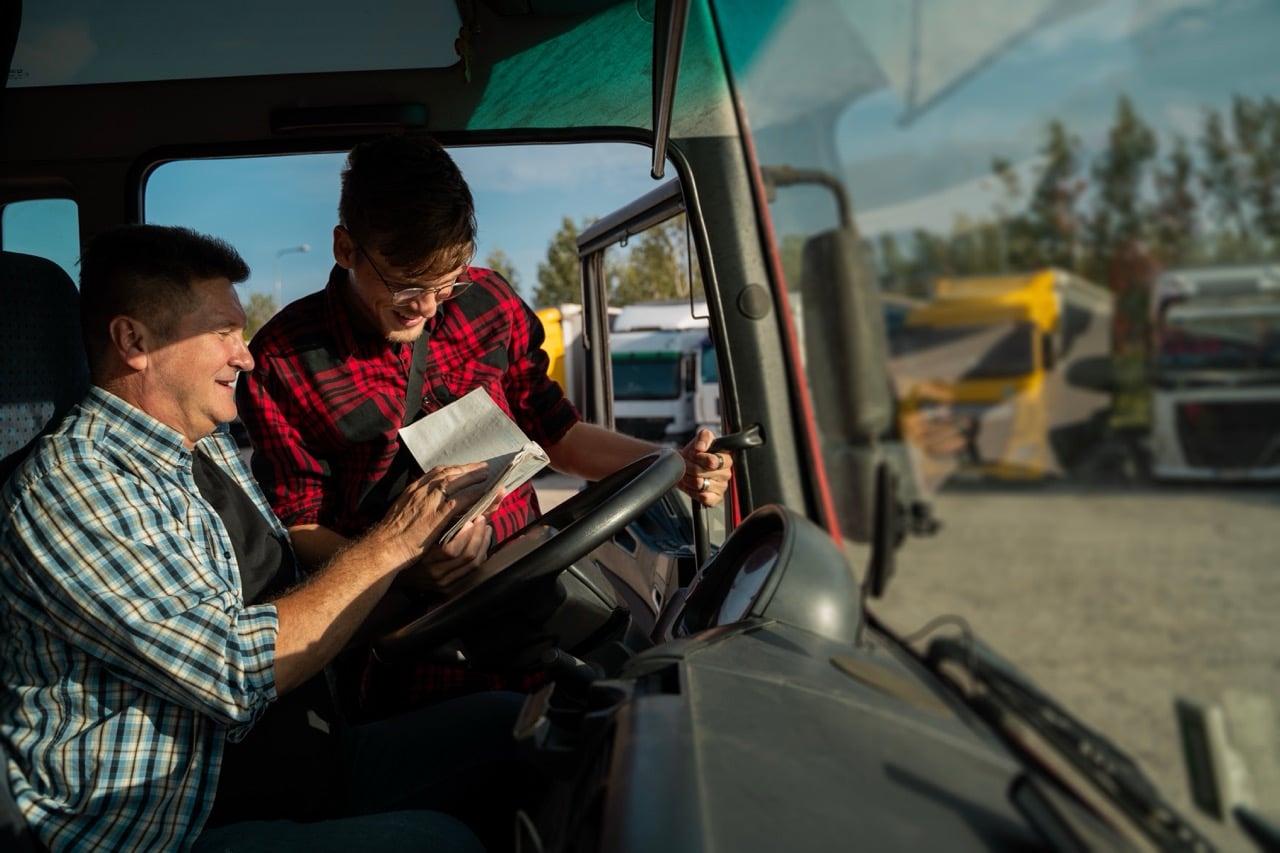 Truck driver and coordinator reviewing load paperwork in a semi cab, showing transportation back office documentation at the source