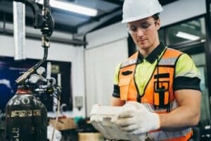 Manufacturing technician in a hard hat and safety vest inspecting a part, highlighting manufacturing document control on the floor
